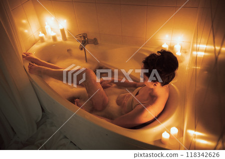 A woman is sitting in a bathtub with candles lit around her. The candles are lit in a way that they create a warm and relaxing atmosphere. A woman is sitting in a bathtub with candles lit around her. The candles are lit in a way that they create a warm and relaxing atmosphere. 118342626
