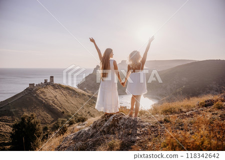 Two women are standing on a hill overlooking the ocean. They are holding hands and looking out at the water. The scene is peaceful and serene, with the sun shining brightly in the background. Two women are standing on a hill overlooking the ocean. They are holding hands and looking out at the water. The scene is peaceful and serene, with the sun shining brightly in the background. 118342642