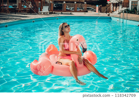 A woman is sitting on a pink inflatable flamingo in a pool. Concept of fun and relaxation, as the woman is enjoying her time in the water 118342709