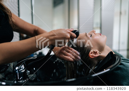 Close-up of unrecognizable female hairdresser washing woman client hair with warm water over sink in hair salon. Blonde female visiting beauty studio for haircut. Concept of fashion and beauty. 118343920