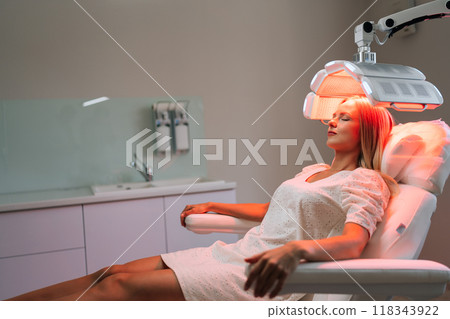 Side view of young woman with blond hair in white uniform having LED light hair photodynamic therapy during hair treatment at medical clinic. Female client having non-invasive type of phototherapy. 118343922