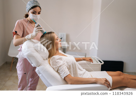Relaxed pretty blonde female client having hair light therapy with modern innovation apparatus. Trichologist in mask and gloves performing hair treatment procedure with spectrum phototherapy device. 118343926