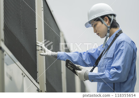 Building maintenance image: A man in work clothes inspecting the outdoor air conditioning unit installed on the roof of a building Building maintenance image: A man in work clothes inspecting the outdoor air conditioning unit installed on the roof of a building 118355654