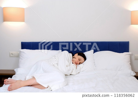 Tired young brunette woman in white bathrobe is sleeping in bed in hotel alone. Tired young brunette woman in white bathrobe is sleeping in bed in hotel alone. 118356062