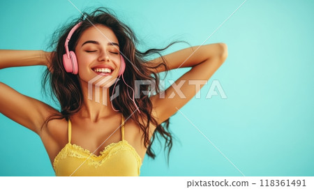 Joyful young woman with medium skin tone, wavy hair, and pink headphones dancing gracefully in a yellow halter top and flowy skirt against a light blue background. Happiness and lifestyle concept 118361491