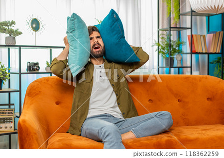 Neighbors repair work, irritated man by loud music at home cover ears with pillows annoyed by noise Neighbors repair work, irritated man by loud music at home cover ears with pillows annoyed by noise 118362259