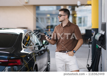 Handsome stylish man waiting for gas station attendant on petrol station. Male driver use his smartphone. Luxury car Handsome stylish man waiting for gas station attendant on petrol station. Male driver use his smartphone. Luxury car 118362449