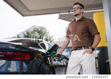 Low angle of concentrated young bearded male driver in sunglasses and casual clothes filling fuel into modern car with gas pump nozzle at station. Luxury car Low angle of concentrated young bearded male driver in sunglasses and casual clothes filling fuel into modern car with gas pump nozzle at station. Luxury car 118362457