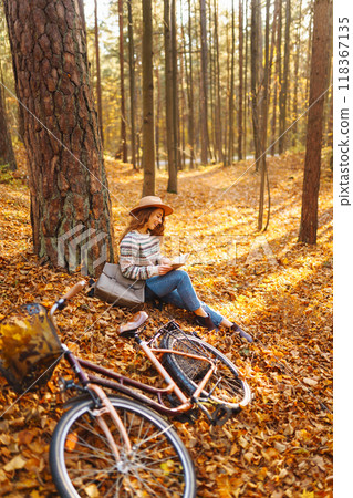 Stylish woman with a book near tree enjoys autumn weather in the park on the background of the bike. 118367135