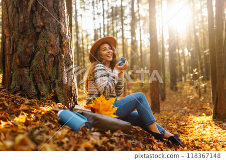 Cute young woman drinks hot drink from thermos and enjoys nature. Autumn landscape. relax concept. Cute young woman drinks hot drink from thermos and enjoys nature. Autumn landscape. relax concept. 118367148