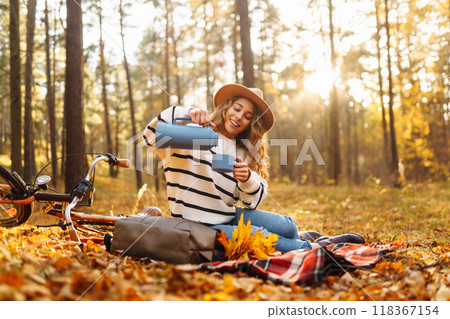 Hiker sitting on picnic blanket, drinking hot drink in autumn forest. Woman resting in nature. 118367154