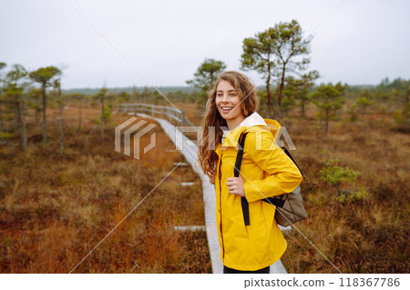 Smiling female traveler in yellow coat walks along wooden path among wild nature. Travel, vacation. 118367786