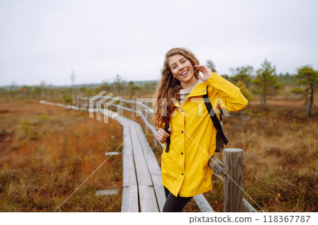Smiling female traveler in yellow coat walks along wooden path among wild nature. Travel, vacation. 118367787