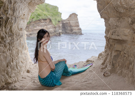 一條年輕的女美人魚坐在一個俯瞰大海的洞穴裡，回頭望去。 118368573