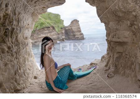 一條年輕的女美人魚坐在一個俯瞰大海的洞穴裡，回頭望去。 118368574