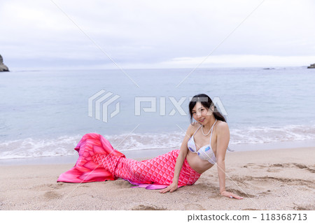 一個年輕的女美人魚坐在沙灘上看著你 118368713
