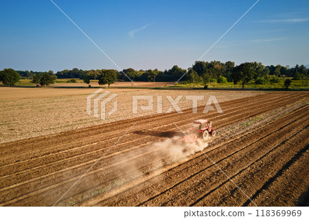Red tractor working in agricultural field, cultivating and plowing dry soil, aerial view. Agribusiness concept. Small farming concept Red tractor working in agricultural field, cultivating and plowing dry soil, aerial view. Agribusiness concept. Small farming concept 118369969