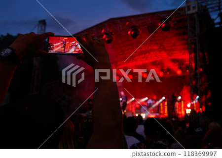 Person standing in front of stage, recording live music concert on smartphone. People crowd near stage enjoys musician band's performance 118369997
