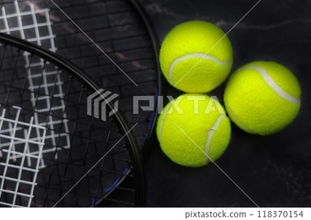 Three yellow tennis balls lie in a wedge in a patch of light, tennis rackets are visible from the side on a dark background, top view. Dark exposure, Low key. High quality photo 118370154