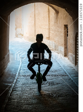 Silhouette of a kid riding a bicycle through a dimly lit tunnel in the narrow streets of the ancient city of Yazd Silhouette of a kid riding a bicycle through a dimly lit tunnel in the narrow streets of the ancient city of Yazd 118371819