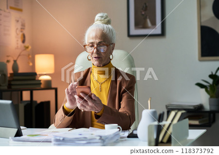 Portrait of Senior Woman Sitting at Office Desk Looking at Phone Portrait of Senior Woman Sitting at Office Desk Looking at Phone 118373792