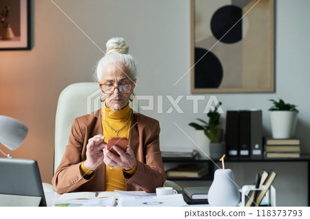 Elderly Woman Checking Phone While Working at Desk Elderly Woman Checking Phone While Working at Desk 118373793