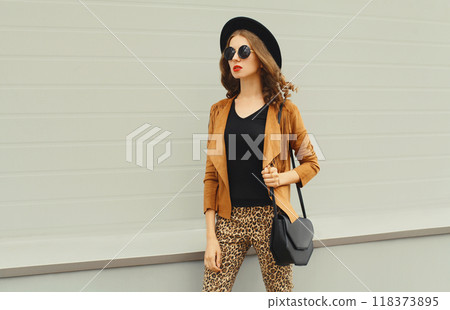 Beautiful stylish young woman posing with handbag in black hat, jacket, glasses on city street 118373895