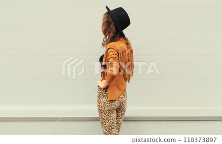 Beautiful stylish young woman posing in black hat, boho style jacket, standing on city street 118373897
