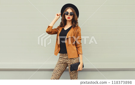 Beautiful stylish young woman posing with handbag in black hat, jacket, glasses on city street 118373898