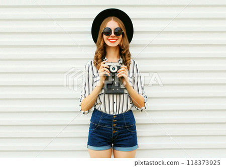 Happy smiling young woman holding camera in black hat, white striped shirt on white wall background Happy smiling young woman holding camera in black hat, white striped shirt on white wall background 118373925