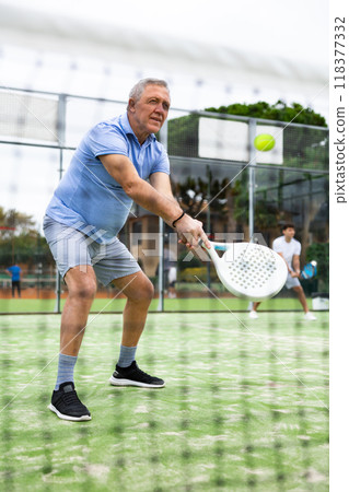 European old man holding playing padel during training in court. View through tennis net European old man holding playing padel during training in court. View through tennis net 118377332