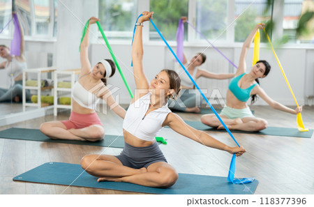 Girl conducts Pilates class for girls sitting with elastic band in hands above head makes body bends 118377396