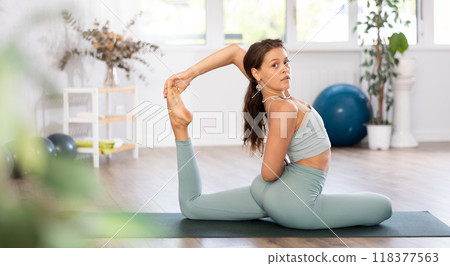 Young woman yoga trainer performing king pigeon pose on black rug 118377563