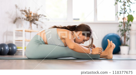 Young woman yoga trainer performing seated forward bend pose on black rug 118377675