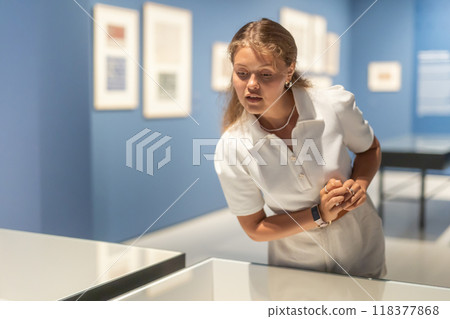 Young female visitor stands near glass display case and examines valuable exhibit Young female visitor stands near glass display case and examines valuable exhibit 118377868