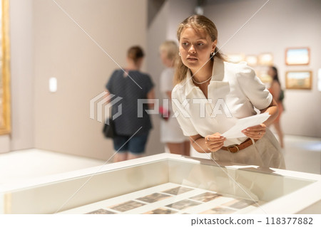 Young female visitor stands near glass display case and examines valuable exhibit Young female visitor stands near glass display case and examines valuable exhibit 118377882