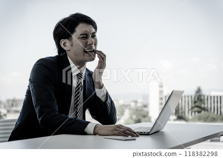 A young Asian male businessman working at a desk is suffering from dry lips, canker sores, tooth decay, and pain. 118382298