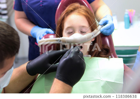 A little girl is being treated for teeth with anesthesia. The girl closed her eyes. Nitrous oxide for pain relief of the child. Treatment of baby teeth. A little girl is being treated for teeth with anesthesia. The girl closed her eyes. Nitrous oxide for pain relief of the child. Treatment of baby teeth. 118386638