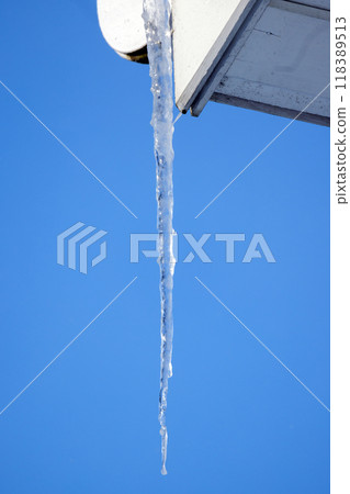 Transparent icicle hangs on a white gutter of rural house Transparent icicle hangs on a white gutter of rural house 118389513