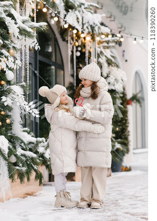 Children girls are standing in front of a Christmas shop window, one of them holding a red candy stick. Festive and joyful 118390260