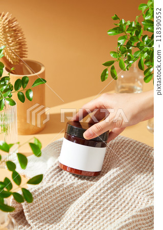 A hand reaches for a brown jar with a blank label, surrounded by green branches and a wooden container, as a template for beauty care product advertising. 118390552