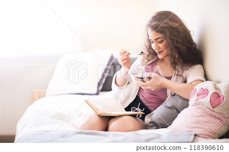 Beautiful young woman with curly hair reading book while eating yogurt bowl with fresh fruits as breakfast. Close up. 118390610