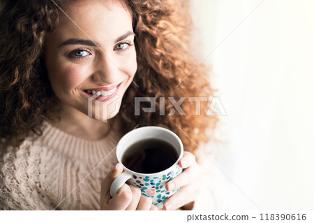 Morning cup of coffee. Beautiful young woman with curly hair sitting on bed in her room, drinking hot coffee. 118390616