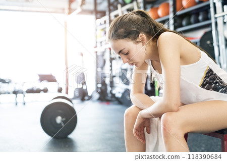 Young fitness girl feeling exhausted and disappointed after hard workout in gym, sitting on bench, breathing. 118390684