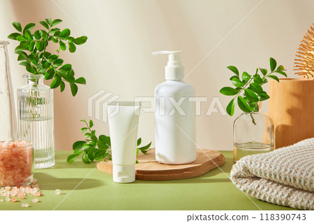 A spa setting features two white beauty products on green backdrop, surrounded by a glass bottle, branches, wooden bowl, pink salt, and white towel, perfect for showcasing skincare or beauty products. 118390743