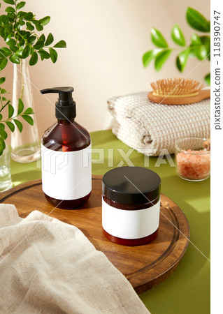 The image showcases an amber glass pump bottle and a black jar with blank labels, surrounded by a wooden cutting board, a white towel, pink salt bowl, and greenery, perfect for beauty product ads. 118390747