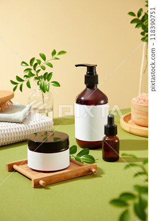 Advertisement beauty products display brown glass pump bottle, dropper bottle, and white cream jar on a green surface, surrounded by green leaves, pink salt, and a white towel. 118390751