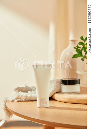The advertisement beauty products features a white tube on a wooden table, a cream jar on a cutting board, a partially visible glass vase with green leaves, and a white linen cloth. The advertisement beauty products features a white tube on a wooden table, a cream jar on a cutting board, a partially visible glass vase with green leaves, and a white linen cloth. 118390882