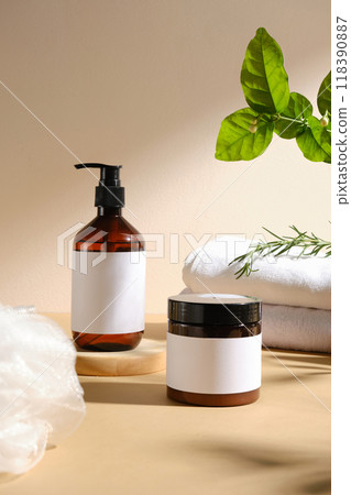 A minimalist still life with brown amber bottle, pump dispenser, cream jar, white towel, bath puff, and leafy sprig, perfect for showcasing beauty products or spa. 118390887