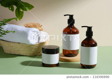 The photo showcases spa and beauty care products, including three brown glass bottles, a jar, a wicker basket, towels, and a hair brush, arranged on a green background with green plants. 118390890